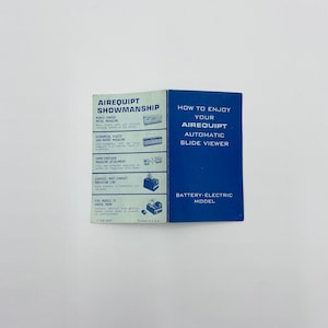 Puede incluir: Un folleto azul y blanco con el título "How to Enjoy Your Airequipt Automatic Slide Viewer" y el texto "Battery-Electric Model". El folleto está doblado por la mitad y muestra las portadas delantera y trasera.