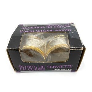 May include: A set of four gold-colored napkin rings in a black box. The box has the text "Brass Napkin Rings Set of 4" and "Ronds de Serviette en Cuivre Ensemble de 4".
