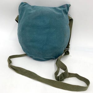 Op de afbeelding: Een teal-kleurige, afgeronde tas met een groene riem. De tas heeft een getextureerde uitstraling en een flap aan de bovenkant. De riem is verstelbaar en gemaakt van duurzaam, geweven materiaal. De tas is ontworpen om over de schouder te worden gedragen.