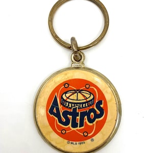 May include: A gold-colored keychain with a round, orange and blue Astros logo. The logo features a baseball stadium with the word "Astros" in a blue font. The keychain also includes the text "MLB 1989" and a copyright symbol.