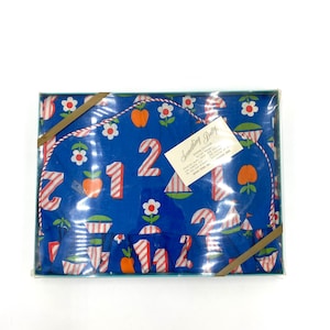 May include: A blue and white fabric with a red and white striped pattern. The fabric has a repeating pattern of numbers, apples, and flowers. The fabric is in a clear plastic box with a white label that says "Something Pretty".