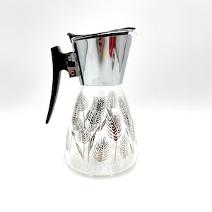 May include: A clear glass pitcher with a silver lid and a black handle. The pitcher has a silver wheat design.