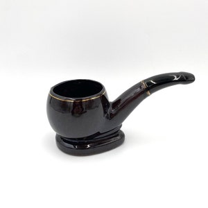 May include: A black ceramic pipe shaped like a traditional tobacco pipe. The pipe has a gold trim around the rim of the bowl.