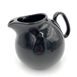 May include: A black ceramic creamer with a rounded body and a handle. The creamer is glossy and reflects light.