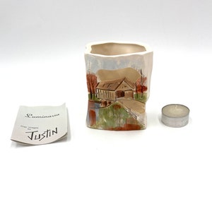 Peut inclure: Un bougeoir en céramique blanche avec un motif découpé représentant un pont couvert et des arbres. Le bougeoir est conçu pour accueillir une bougie chauffe-plat. Le bougeoir est posé sur une surface blanche avec une petite bougie chauffe-plat argentée à côté. Une carte avec le texte "Luminaria Clay Images by Justin" est également présente sur l'image.