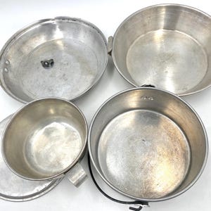 Op de afbeelding: Een set vintage aluminium kampeerkookgerei, waaronder een pot met een handvat, een beker met een handvat en twee pannen. Het metaal heeft een versleten uiterlijk. Ideaal voor buiten koken en kamperen.