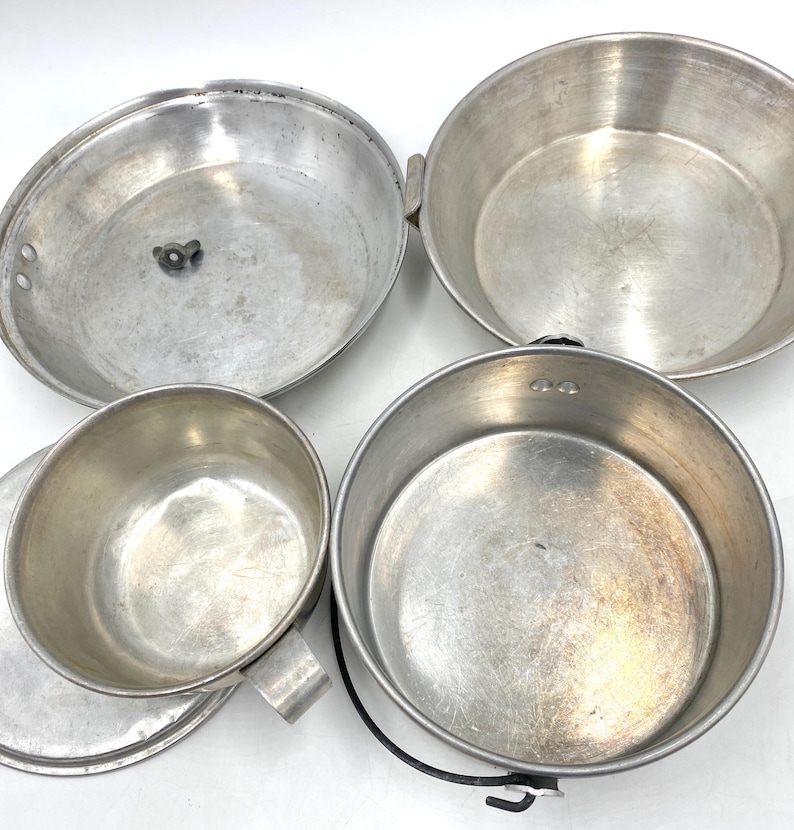 Op de afbeelding: Een set vintage aluminium kampeerkookgerei, waaronder een pot met een handvat, een beker met een handvat en twee pannen. Het metaal heeft een versleten uiterlijk. Ideaal voor buiten koken en kamperen.