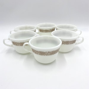 Puede incluir: Seis tazas de té blancas con un diseño floral marrón en el borde dispuestas en círculo. Cada taza tiene un asa y una forma simple y elegante. Las tazas están sobre un fondo blanco liso, resaltando su diseño.