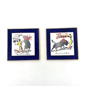 May include: Two framed ceramic tiles with a blue background and gold trim. Each tile features a black and white illustration of a bullfight scene with a matador and a bull. The text "Recuerdo de Rota" is printed on each tile.