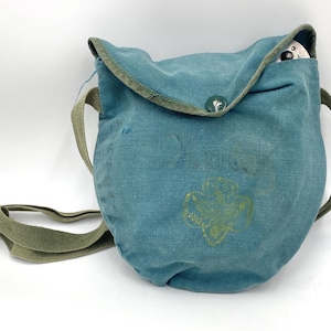 Op de afbeelding: Teal canvas schoudertas met een groene band en afwerking. De tas heeft een Girl Scout-embleem en een knoopsluiting. Een klein wit voorwerp is zichtbaar in de tas.