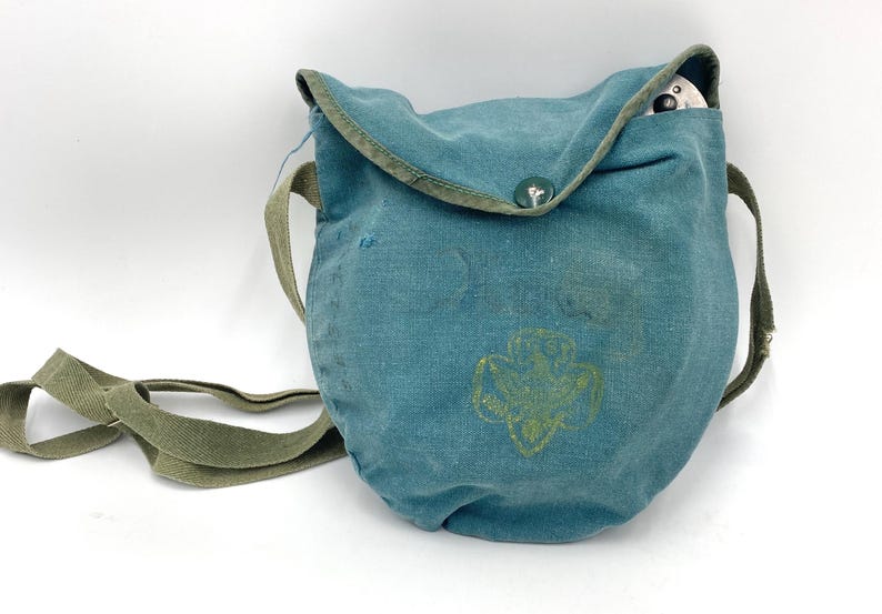 Op de afbeelding: Teal canvas schoudertas met een groene band en afwerking. De tas heeft een Girl Scout-embleem en een knoopsluiting. Een klein wit voorwerp is zichtbaar in de tas.