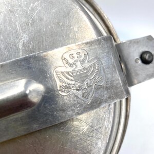 Op de afbeelding: Een vintage, zilverkleurige metalen kampeermesset. De set heeft een deksel, een handvat en het reli&euml;f van het Girl Scouts of the USA-embleem met de letters "GS" en een adelaar. Het metaal vertoont tekenen van slijtage.