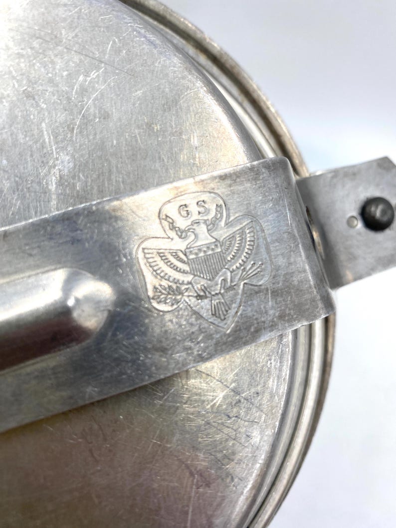 Op de afbeelding: Een vintage, zilverkleurige metalen kampeermesset. De set heeft een deksel, een handvat en het reli&euml;f van het Girl Scouts of the USA-embleem met de letters "GS" en een adelaar. Het metaal vertoont tekenen van slijtage.
