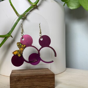 May include: Plum-colored Mickey Mouse-shaped earrings with gold-colored ear wires. One earring features a gold glitter bow. The earrings are displayed on a clear acrylic stand with a wooden base. The background is a white vase and green plant.