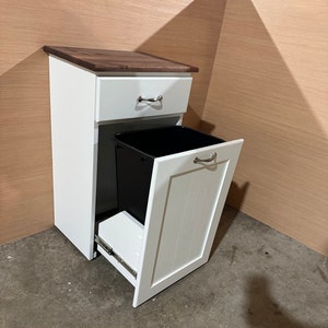 May include: A white wooden trash cabinet with a dark brown countertop. The cabinet has a drawer with a silver handle and a hinged door that opens to reveal a black trash bin. The door also has a silver handle.