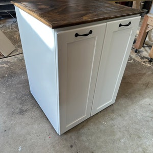 May include: White double-door cabinet with a wood top and black handles. The cabinet is standing on a concrete floor.