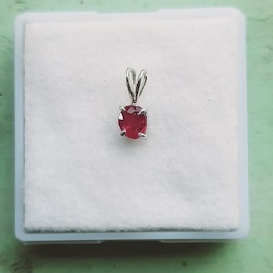 May include: A pendant featuring a small, oval-cut, deep red gemstone secured in a four-prong silver setting. The piece includes a simple silver bail at the top for attachment to a chain, presented against a plain, white, textured square background.