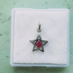 Puede incluir: Un colgante en forma de estrella de tono plateado con una piedra preciosa de color rojo intenso y corte ovalado en el centro. La estrella de metal tiene un acabado texturizado ligeramente desgastado y una pequeña anilla integrada.