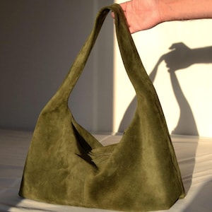 Può includere: Borsa a spalla in camoscio verde oliva con una lunga tracolla. La borsa ha un design morbido e ampio e un'estetica semplice e minimalista. La borsa è tenuta da una mano.