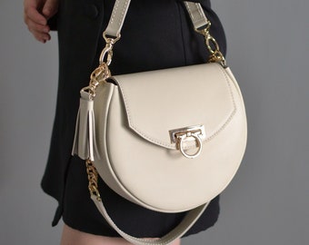 Könnte beinhalten: Eine cremefarbene Ledertasche mit einer goldenen Kettenkette und einem goldenen Quastenanhänger. Die Tasche hat eine Halbkreisform und einen goldenen Verschluss.