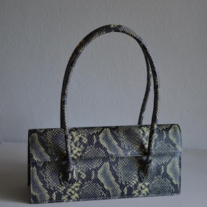 Könnte beinhalten: Eine rechteckige Handtasche mit einem schwarz-gelben Schlangenledermuster. Die Tasche verfügt über einen strukturierten Klappverschluss und zwei dünne, passende Tragegriffe im Schlangenledermuster, die an der Vorderseite befestigt sind.