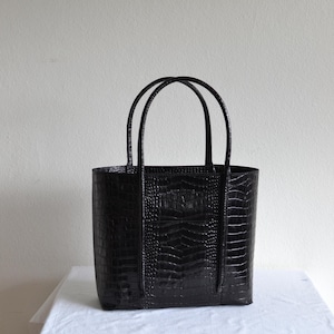 Può includere: Una borsa tote nera con una texture a pelle di coccodrillo. La borsa ha due manici e una forma rettangolare. La borsa è su una superficie bianca.