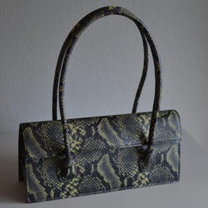 Könnte beinhalten: Eine rechteckige Handtasche mit einem schwarz-gelben Schlangenledermuster. Die Tasche hat einen strukturierten, horizontalen Körper mit Klappverschluss und zwei hohe, dünne, passende Tragegriffe mit Schlangenledermuster an der Vorderseite.