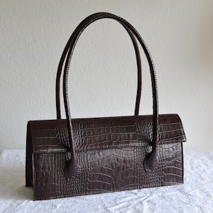 May include: A rectangular dark brown handbag featuring a textured crocodile skin pattern. The bag has a structured horizontal body with a flap closure and two long, thin, matching handles attached to the front and back panels of the exterior.
