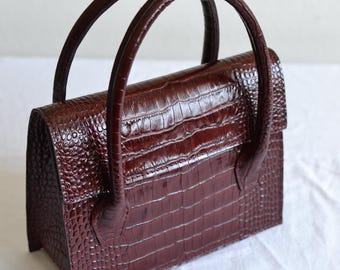 May include: A burgundy-colored handbag with a crocodile skin texture. The bag has two rounded handles and a rectangular shape with a flap closure. The bag is made of a shiny, textured material.