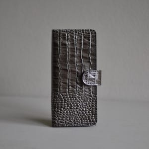 May include: A dark grey rectangular wallet featuring a textured crocodile skin pattern. The wallet has a vertical orientation with a small, matching textured tab closure on the right side. The surface displays a mix of large and small scale patterns.