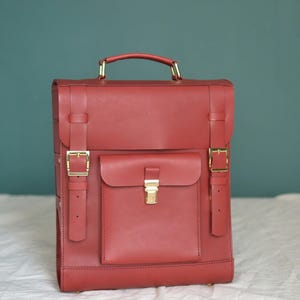 May include: A red leather backpack with a large front pocket and gold buckles. The backpack has a top handle and adjustable straps.