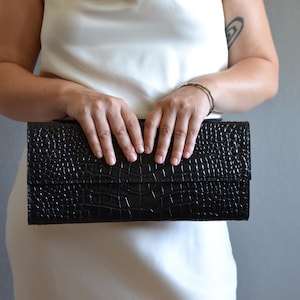 Könnte beinhalten: Eine rechteckige schwarze Clutch mit einer strukturierten, geprägten Krokodilleder-Optik. Die Tasche hat eine längliche, strukturierte Form mit einem horizontalen Klappverschluss, der sich über die gesamte Vorderseite erstreckt.