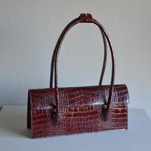 May include: A rectangular handbag in a deep burgundy colour with a textured crocodile skin pattern. The bag features a structured flap closure and two long, thin shoulder straps that are joined at the top by a small, matching rectangular tab.