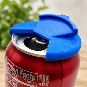 May include: A bright blue plastic beverage can cover featuring a three-part segmented design that snaps onto the top rim of a red aluminum soda can. The cover partially obscures the top opening while leaving the pull tab mechanism visible.