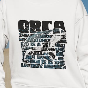 Könnte beinhalten: Weißer Pullover mit einem schwarzen Grafikdruck eines Schwertwals. Der Text lautet "ORCA Das größte Mitglied der Familie der ozeanischen Delfine, das gleichzeitig das größte Mitglied ist".