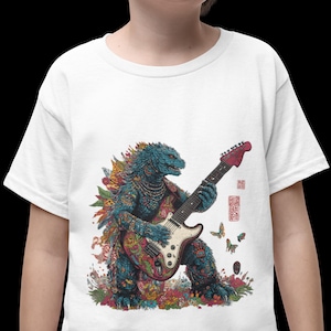 以下が含まれることがあります： 白いTシャツに、エレキギターを演奏するゴジラのカラフルなイラスト。青とターコイズ色のゴジラが、花や蝶に囲まれて描かれており、鮮やかなイメージを創出しています。