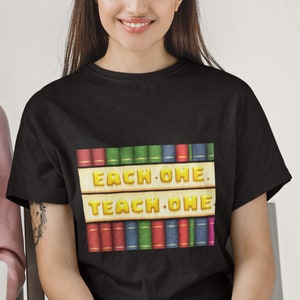 以下が含まれることがあります： 黄色い文字で「EACH ONE TEACH ONE」と書かれた本棚のグラフィックがプリントされた黒いTシャツです。