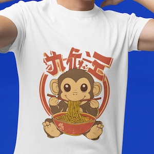 以下が含まれることがあります： 赤い丼から箸でラーメンを食べる漫画の猿が描かれた白いTシャツ。デザインには赤い円の中に日本語のテキストが含まれています。猿は茶色です。