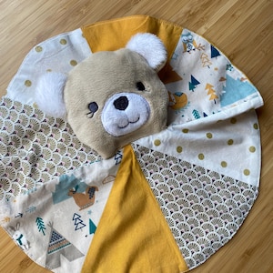 Puede incluir: Una manta de seguridad para bebés con una cabeza de oso de peluche y orejas blancas. La manta circular está hecha de paneles de tela en amarillo, crema y verde, con patrones de animales del bosque y geométricos. La manta está diseñada para la comodidad.