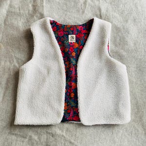 Pode incluir: Um colete sem mangas com um exterior de lã branca texturizada e um forro interior contrastante com um padrão floral vibrante em tons de vermelho, laranja, azul e verde. Uma pequena etiqueta retangular está centrada na linha do pescoço.