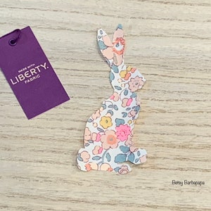 Peut inclure: Une découpe de lapin en tissu blanc avec un motif floral rose, jaune et bleu. Le lapin est assis sur une surface en bois. Le lapin a une étiquette attachée à lui qui dit "Fabriqué avec du tissu Liberty."
