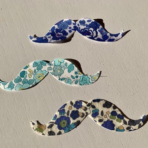 Peut inclure: Trois moustaches en tissu de formes différentes avec des motifs floraux. La moustache du haut est bleue et blanche, la moustache du milieu est bleu clair et blanche, et la moustache du bas est bleu foncé et blanche.