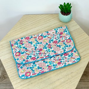 Puede incluir: Un embrague de estampado floral azul y blanco con ribete turquesa. El embrague tiene un cierre de botón único.