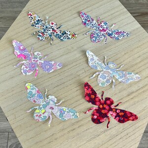 Può includere: Sei sagome di carta a forma di ape colorate con motivi floreali. Le api sono disposte a semicerchio su una superficie di legno chiaro.