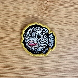 May include: Embroidered patch featuring a whimsical fish design. The patch has a yellow border and showcases a detailed fish with a patterned body in shades of white and black, with a pink mouth and large eyes. This patch is suitable for clothing or accessories.