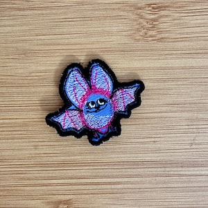 May include: Embroidered patch featuring a cartoon bat with blue and pink wings and body, outlined in black. The bat has a smiling face with black eyes. The patch is likely for clothing or accessories.