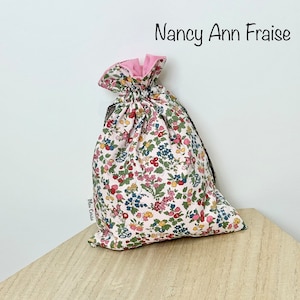 Peut inclure: Un sac à cordon rose et blanc avec un imprimé floral. Le sac a une doublure rose et un cordon de serrage noir. Le sac est posé sur une surface en bois. Le texte "Nancy Ann Fraise" est écrit en noir au-dessus du sac. Le texte "Elu Cruse" est écrit en noir sur le côté du sac.