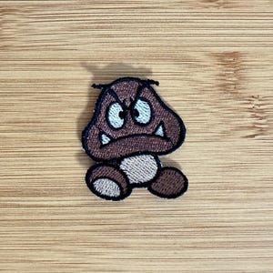 May include: An embroidered patch featuring a brown, mushroom-shaped character with a stern expression, white eyes, and two small white fangs. The character has a rounded brown top, a light tan lower body, and two small brown feet, all outlined in black thread.