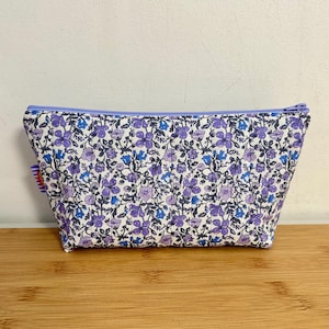 Pode incluir: Uma bolsa de cosméticos com estampa floral e zíper lavanda. A bolsa apresenta um padrão de flores roxas e azuis em um fundo branco. A bolsa é retangular e fica em uma superfície de madeira.