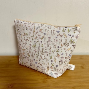 Op de afbeelding: Een witte make-uptas met een bloemen- en dierenpatroon. De tas heeft een gouden rits en een klein wit label. Het patroon bevat vogels, bloemen en kleine dieren in blauwe, groene en paarse tinten.